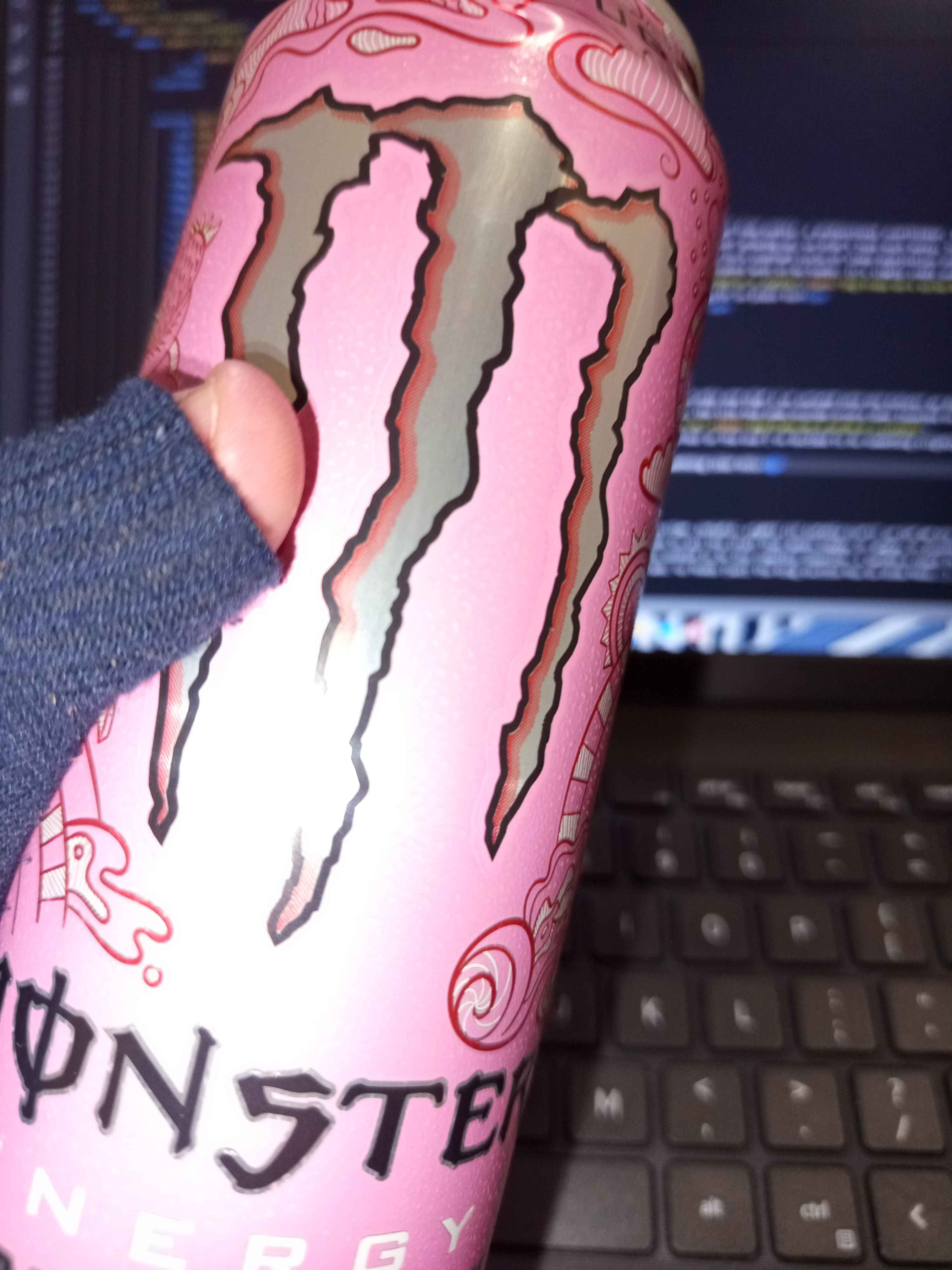 Hand holding Monster Zero Ultra Strawberry Dreams can; laptop with VS Code in background.